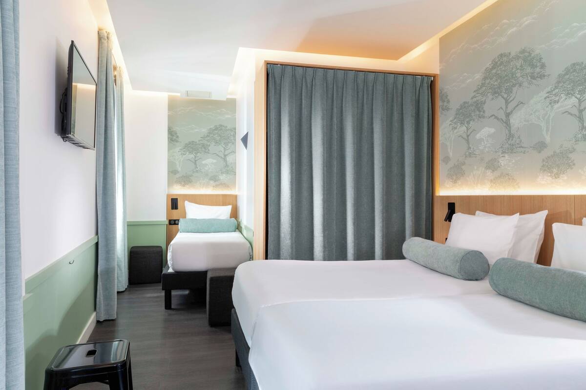 A hotel room with two beds, a small stool, a wall-mounted TV, and a large window with curtains.