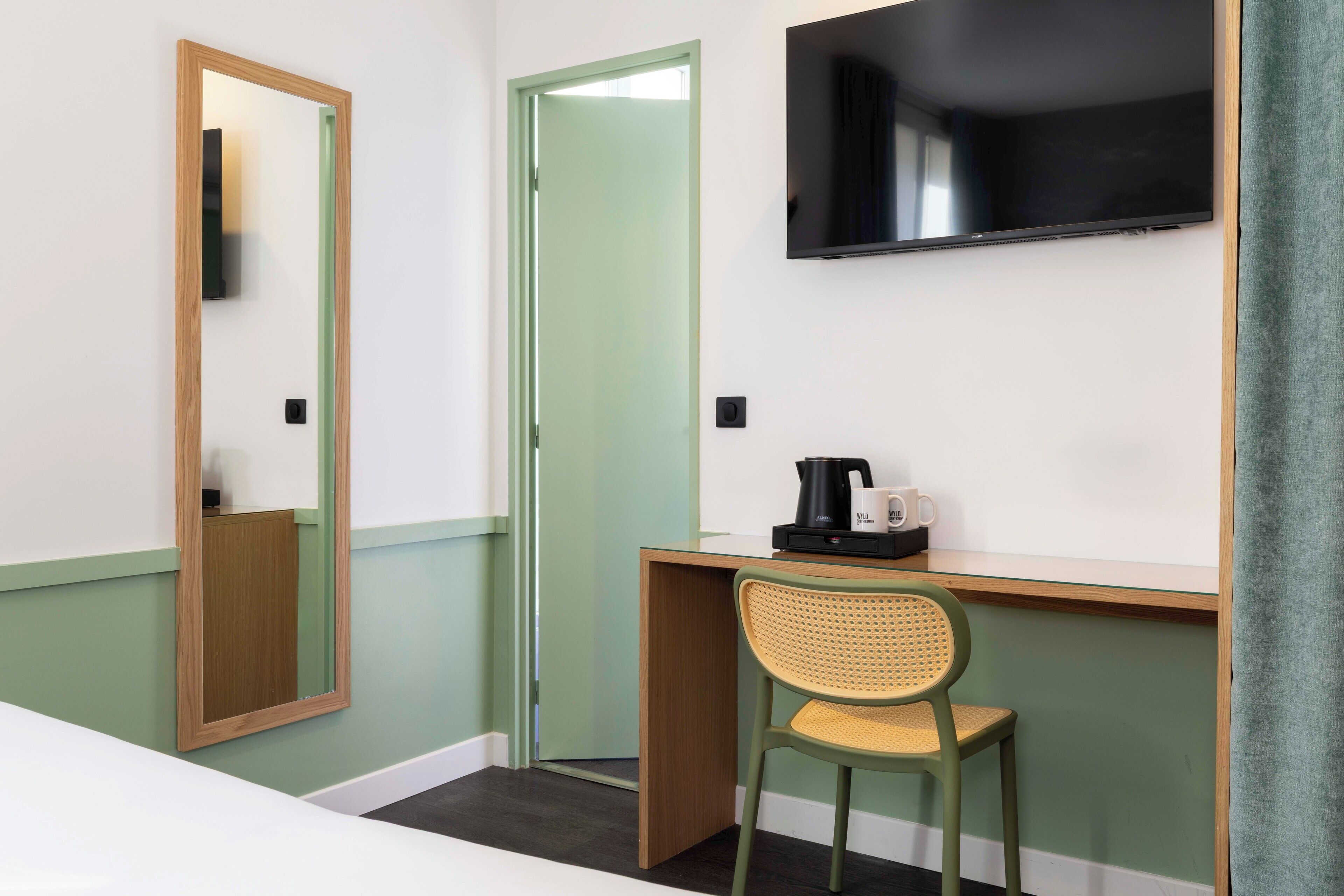 Habitación de hotel con un escritorio de madera, una silla verde, un televisor de pantalla plana montado en la pared y una puerta verde.