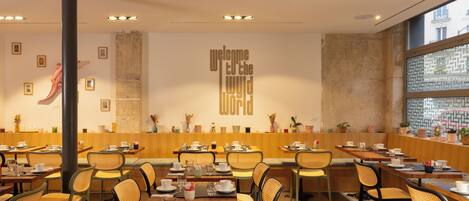 Un restaurant moderne avec des tables en bois et des chaises en osier, une grande colonne et une pancarte sur le mur indiquant : « Bienvenue dans le monde ».