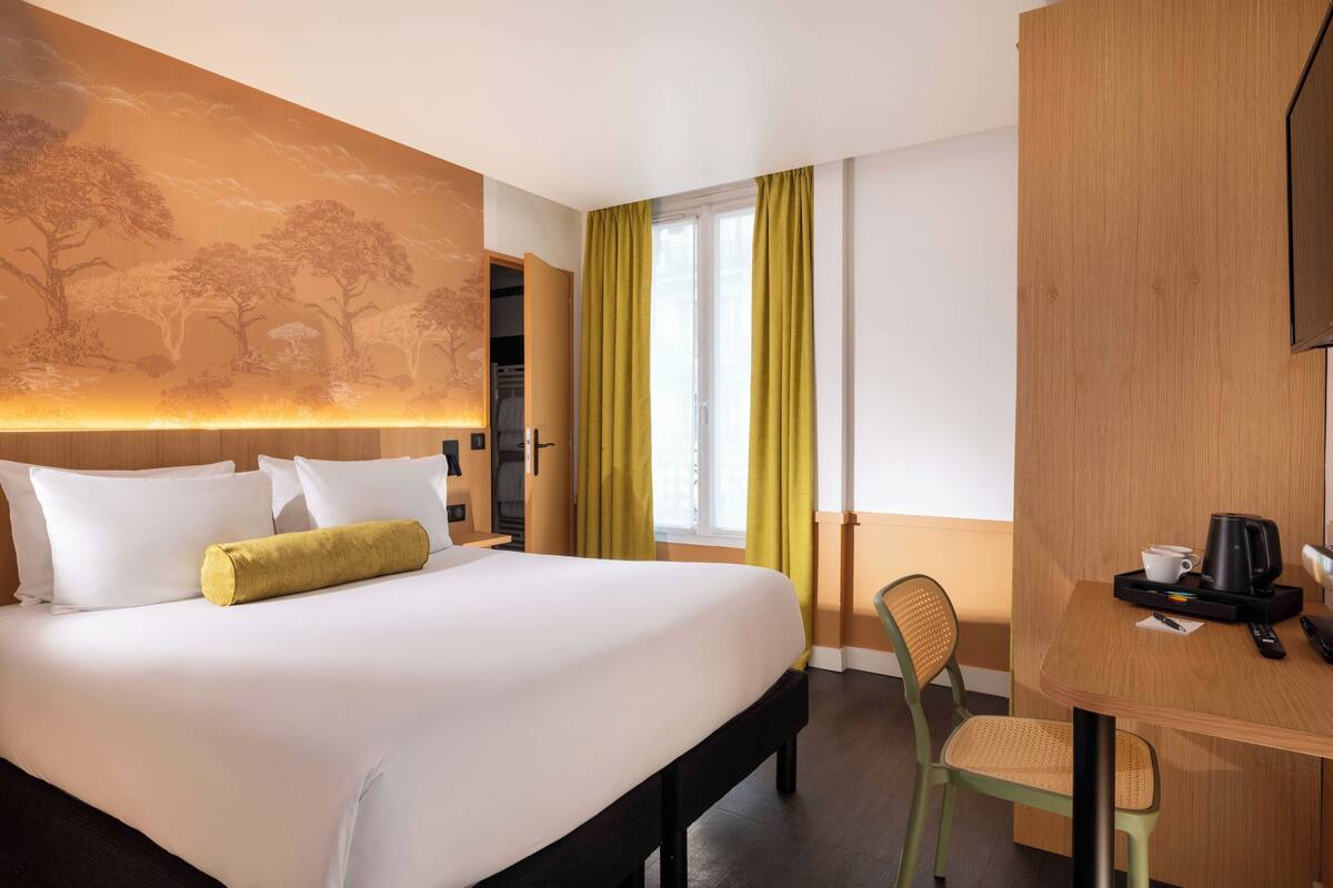 A hotel room with a large bed, a desk with a chair, a coffee maker, and a window with curtains.