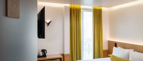 Habitación de hotel con cama, escritorio con cafetera, una silla y ventana con cortinas amarillas.