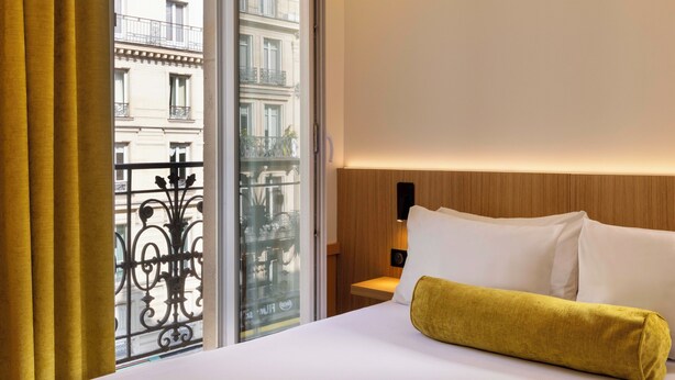 Una habitación de hotel con una cama, un ventanal con cortinas y vistas a edificios.