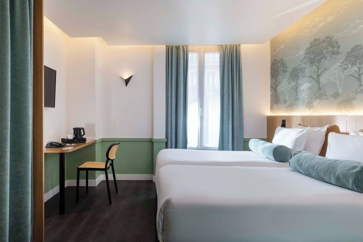 A hotel room with two beds, a desk with a chair, a wall-mounted light, and a window with curtains.