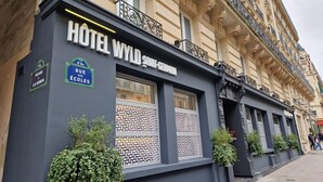 Un hotel con fachada moderna, llamado "HOTEL WYLD SAINT-GERMAIN", presenta grandes ventanales con persianas ornamentales, un cartel que indica "5º Arreglo, Rue DE ÉCOLES", y un balcón con barandillas decorativas.