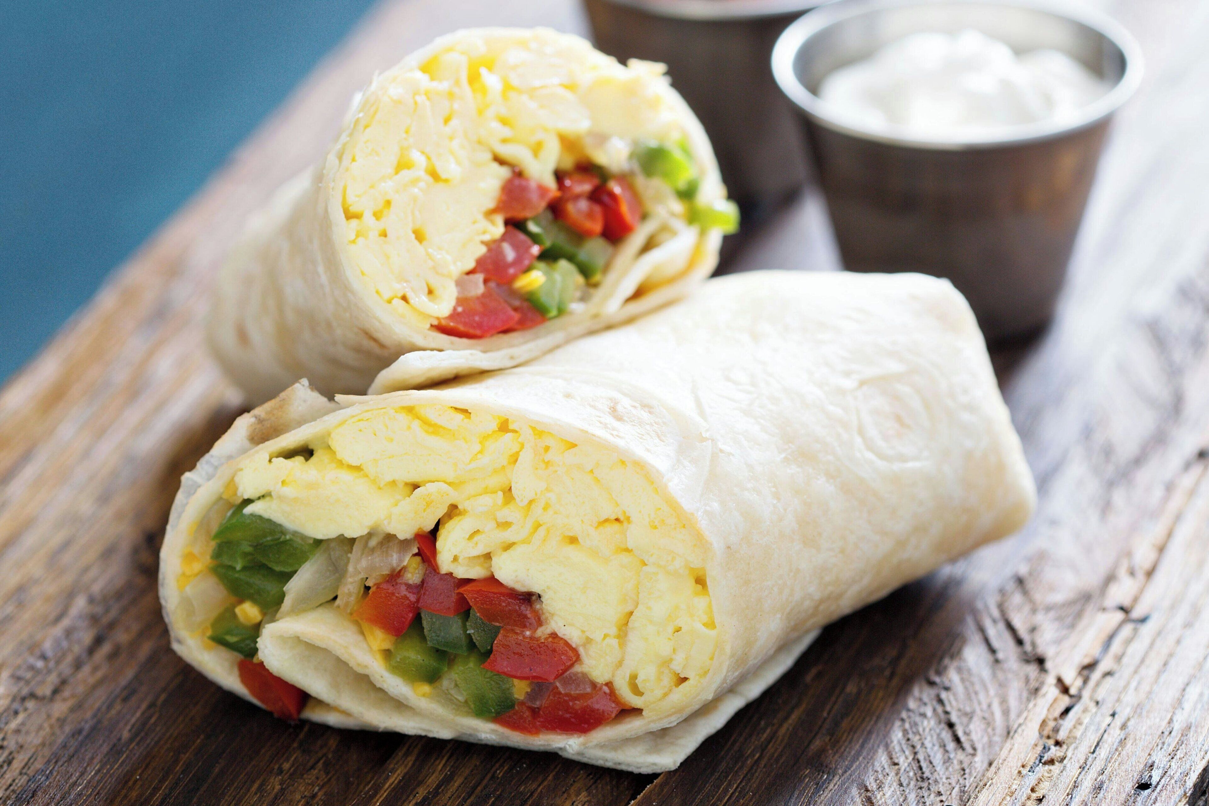 A burrito cut in half on a wooden surface with a metal cup of sour cream in the background.