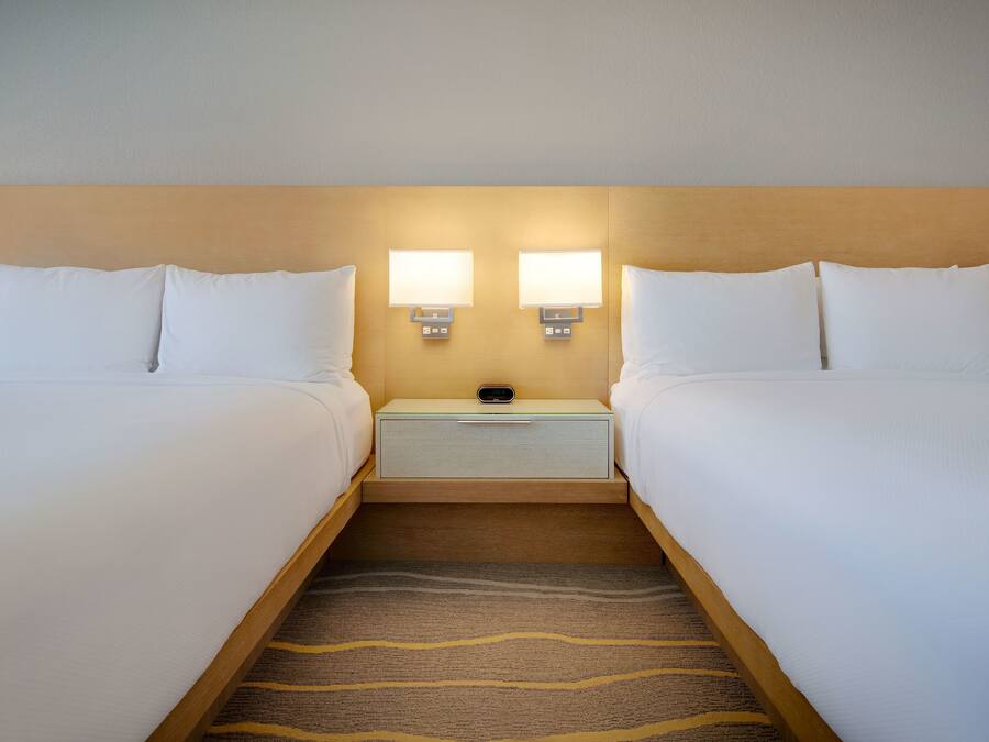 Quarto de hotel com duas camas, uma mesa de cabeceira com duas luminárias fixadas na parede e uma gaveta pequena.