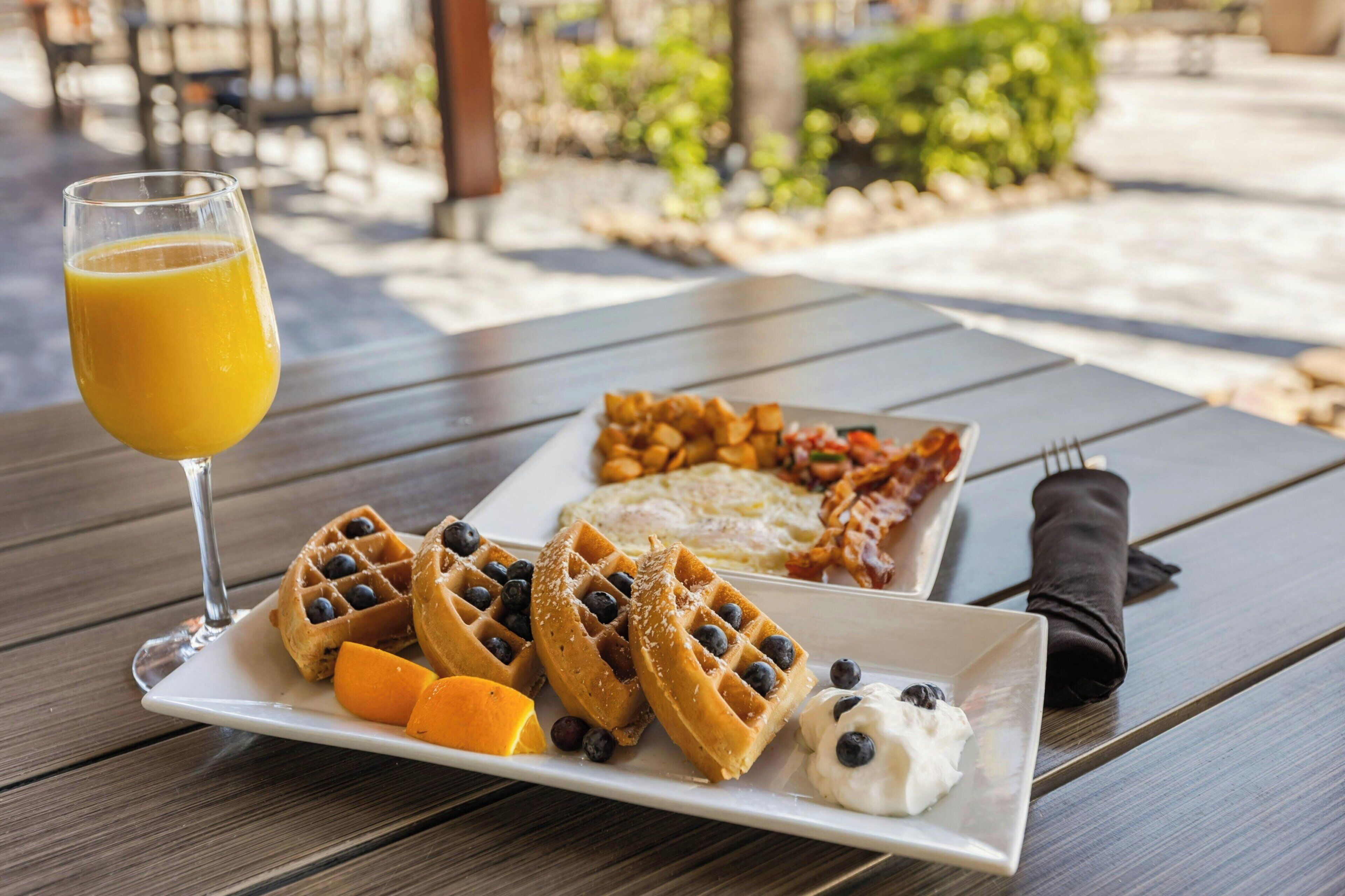 Prato de café da manhã com waffles, amoras, rodelas de laranja e um copo de suco de laranja sobre uma mesa de madeira.