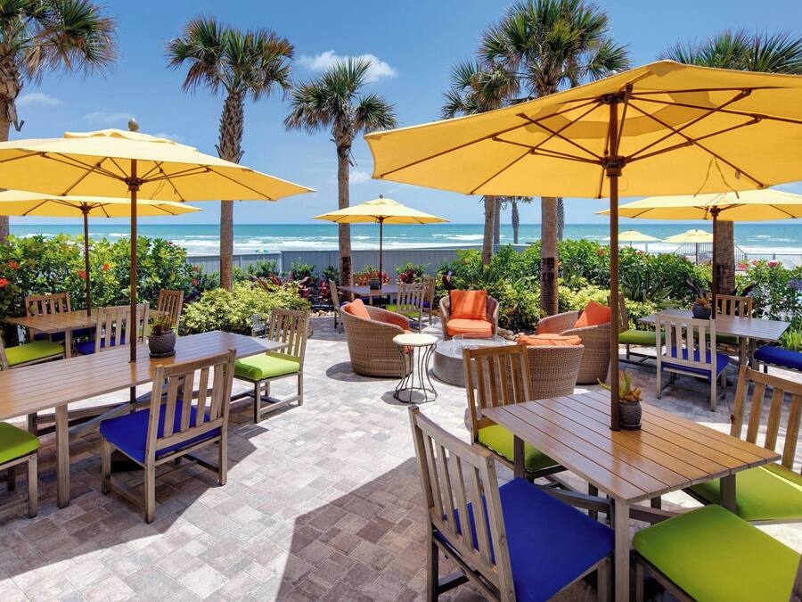 An outdoor seating area with wooden tables and chairs, yellow umbrellas, and a view of the ocean.