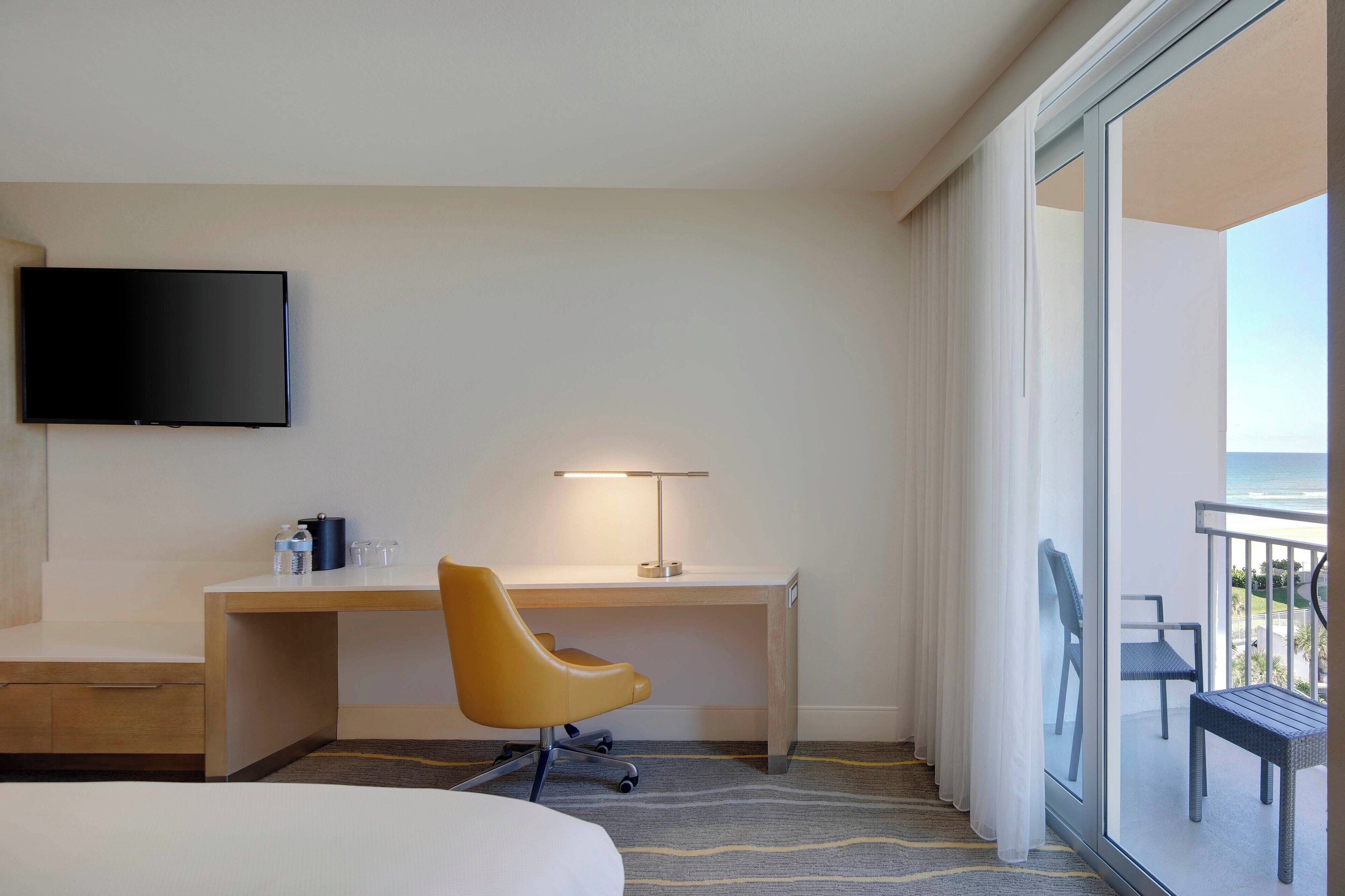 A hotel room with a bed, a desk with a chair, a TV, and a balcony with a view of the ocean.