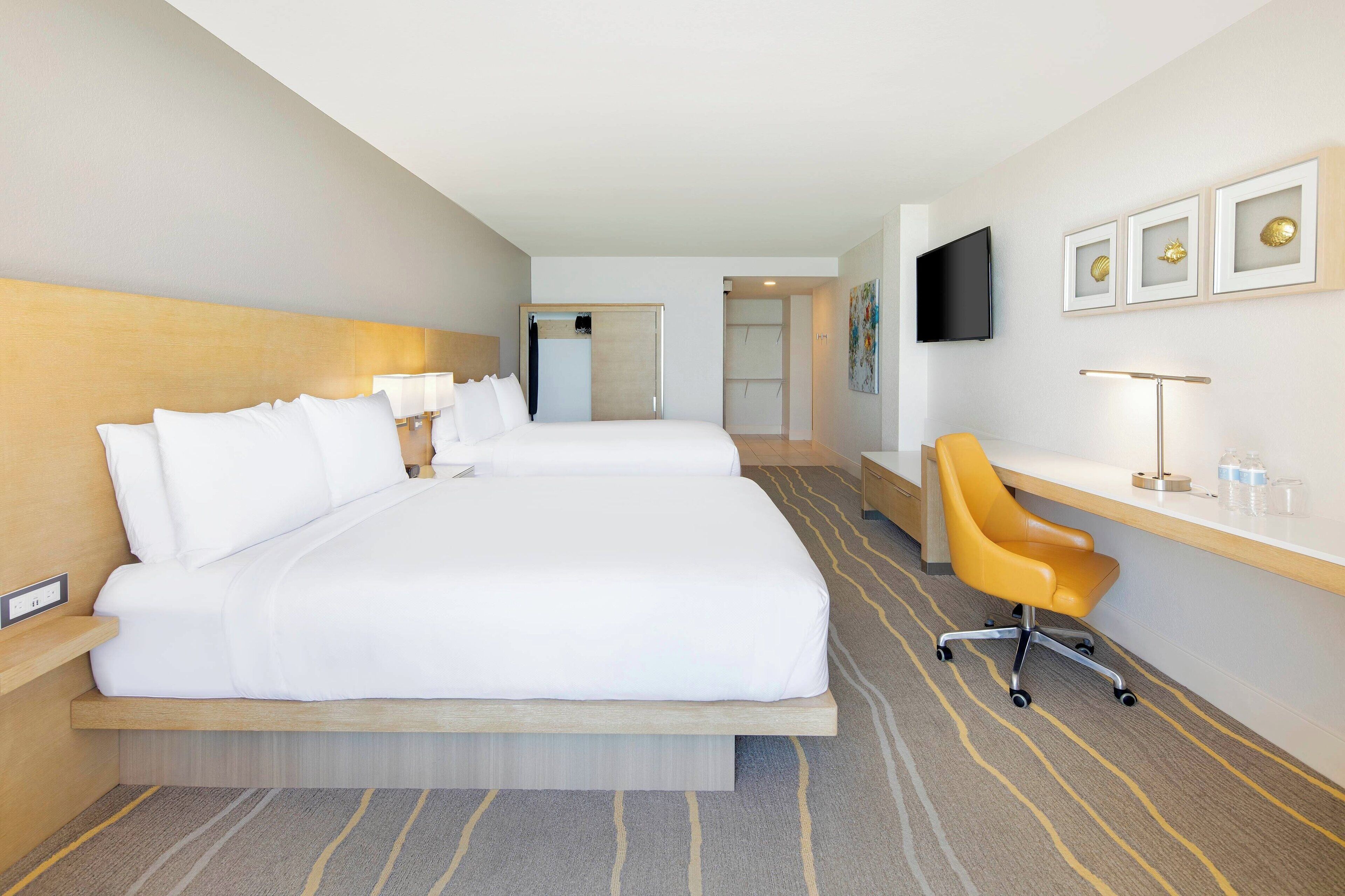 Une chambre d’hôtel avec deux lits, un bureau, une chaise, une télévision et des œuvres d’art encadrées accrochées au mur.