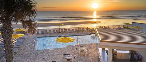 A poolside area with lounge chairs and umbrellas, overlooking a beach and ocean at sunset.