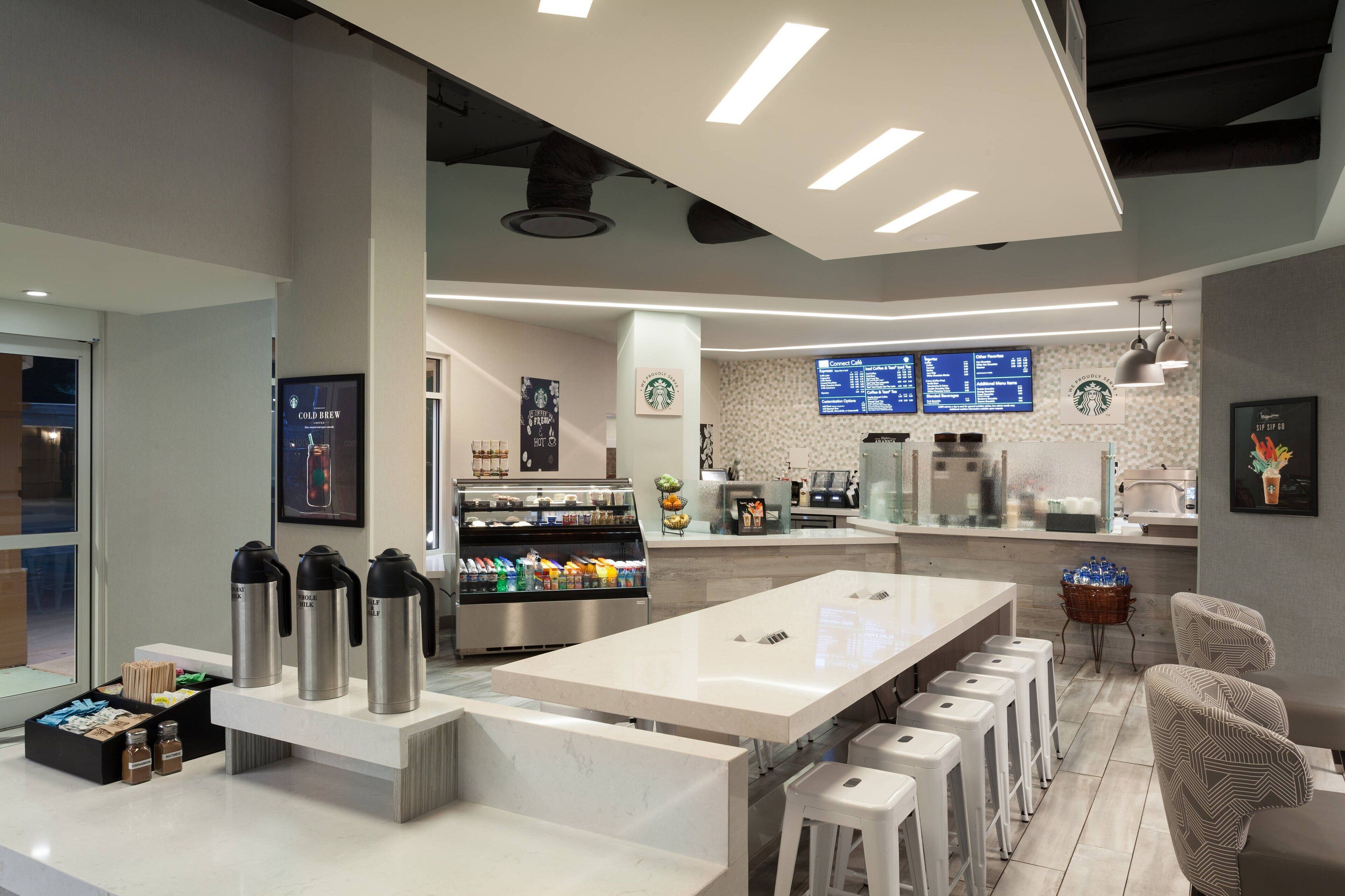 Un interior de cafetería moderna con una barra larga, zona de estar y una pantalla de menú.