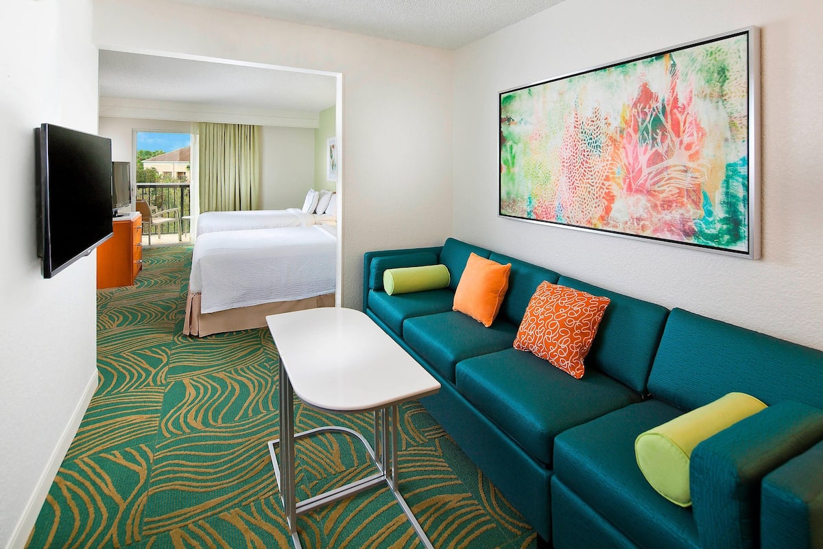Habitación de hotel con alfombra verde estampada, sofá color turquesa con cojines naranja y amarillos, un televisor de pantalla plana y un cuadro abstracto de gran tamaño en la pared.