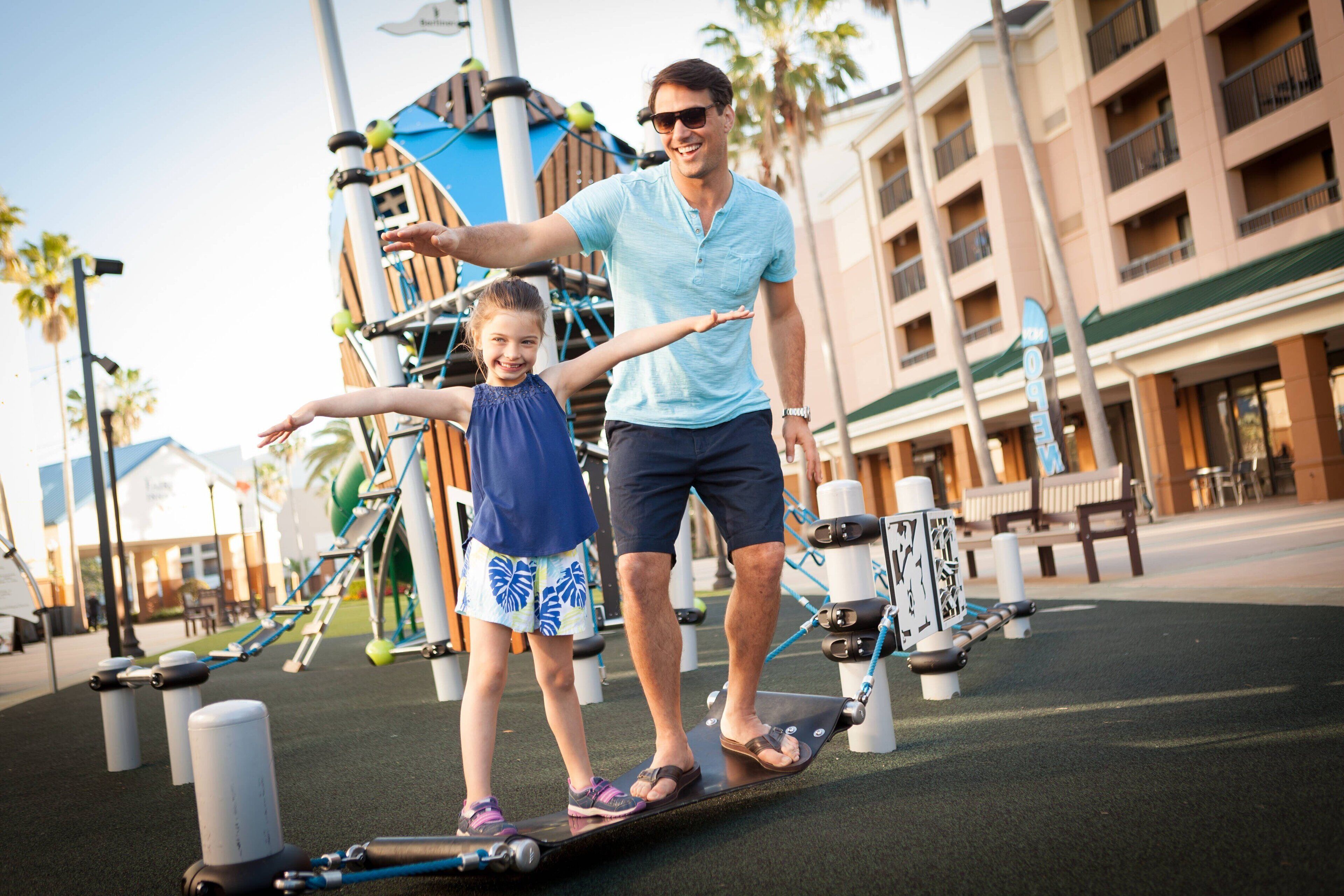 A man and a child on a seesaw at an outdoor playground with exercise equipment and palm trees in the background.