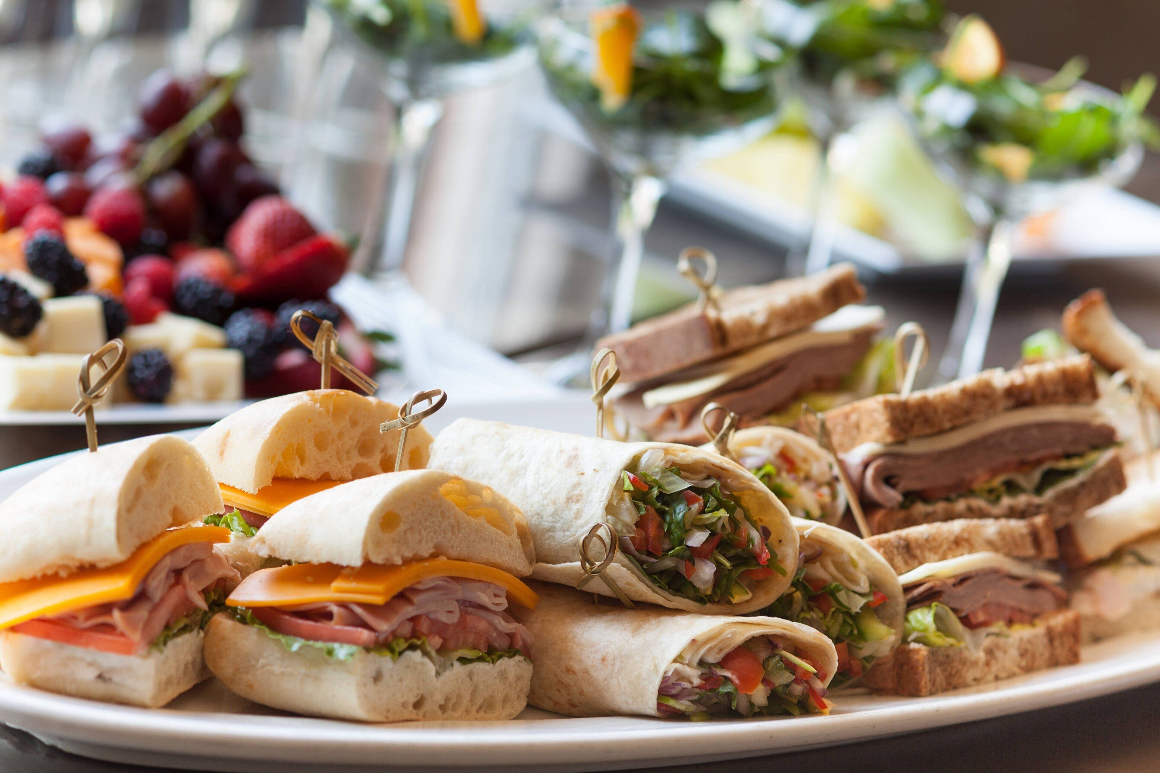 Un plato de sándwiches con diversos rellenos, incluyendo carnes y quesos, y un bol de frutos rojos al fondo.