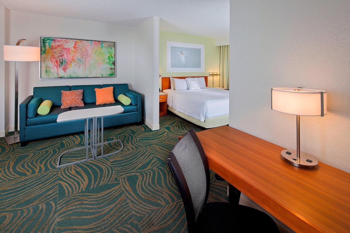 Habitación de hotel con alfombra verde estampada, sofá color turquesa con cojines naranja y amarillos, escritorio de madera, silla negra, cama con ropa blanca y dos lámparas fijadas a la pared.