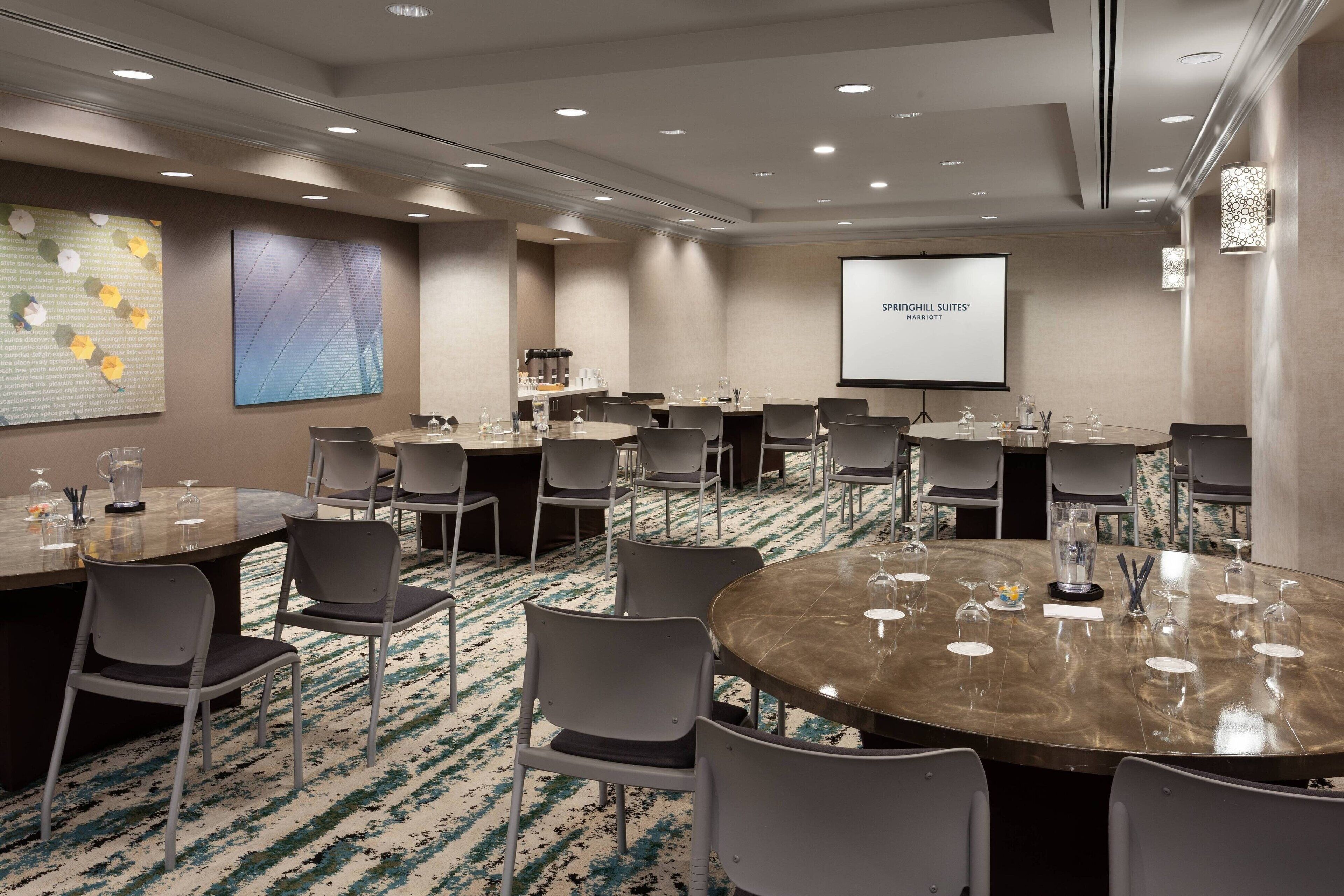Une salle de conférence équipée de plusieurs tables et chaises, d’un écran de projection affichant le texte « SPRINGHILL SUITES® MARLBOROUGH », et d’une machine à café.