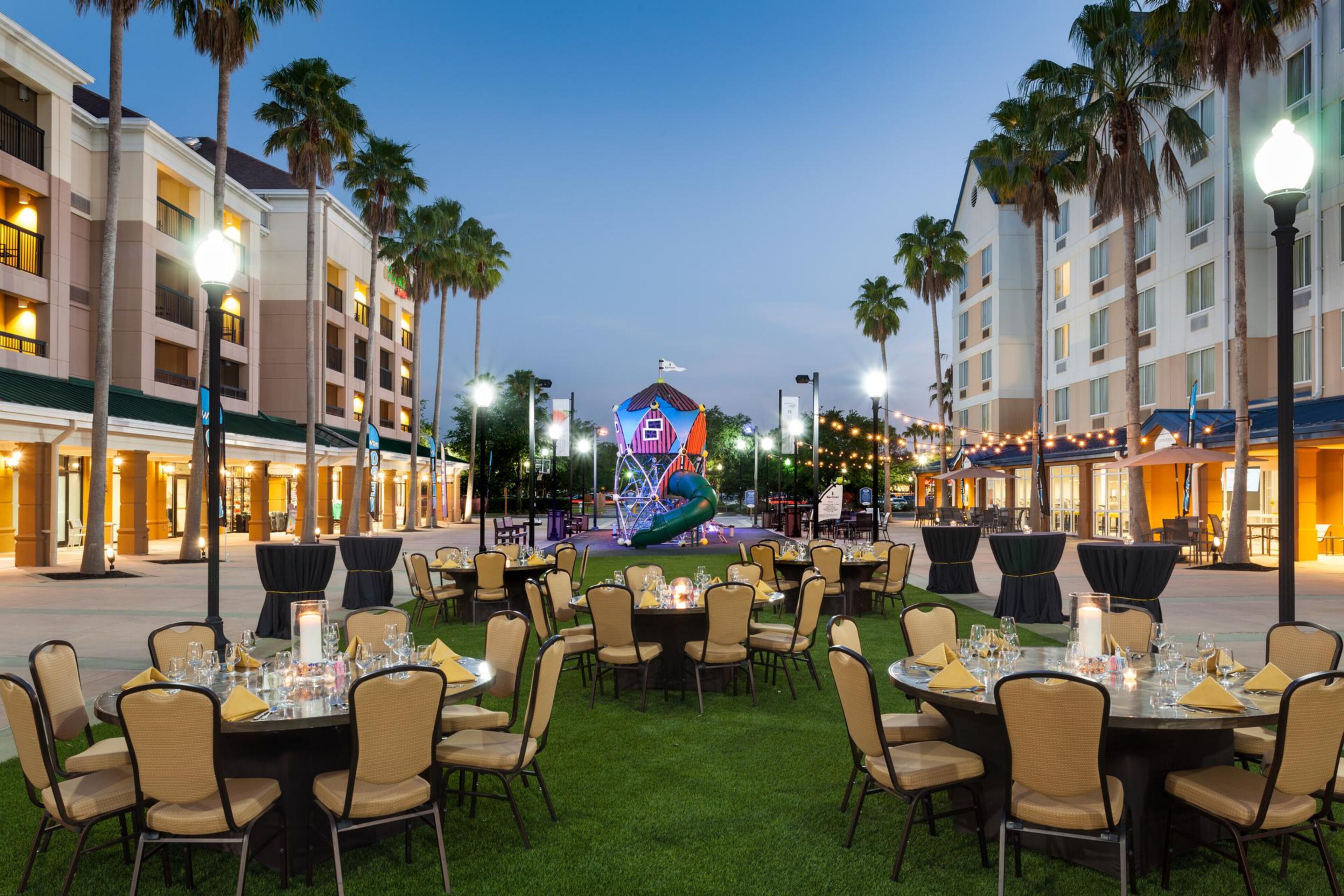 An outdoor dining area with round tables and chairs, surrounded by palm trees and a building with balconies.