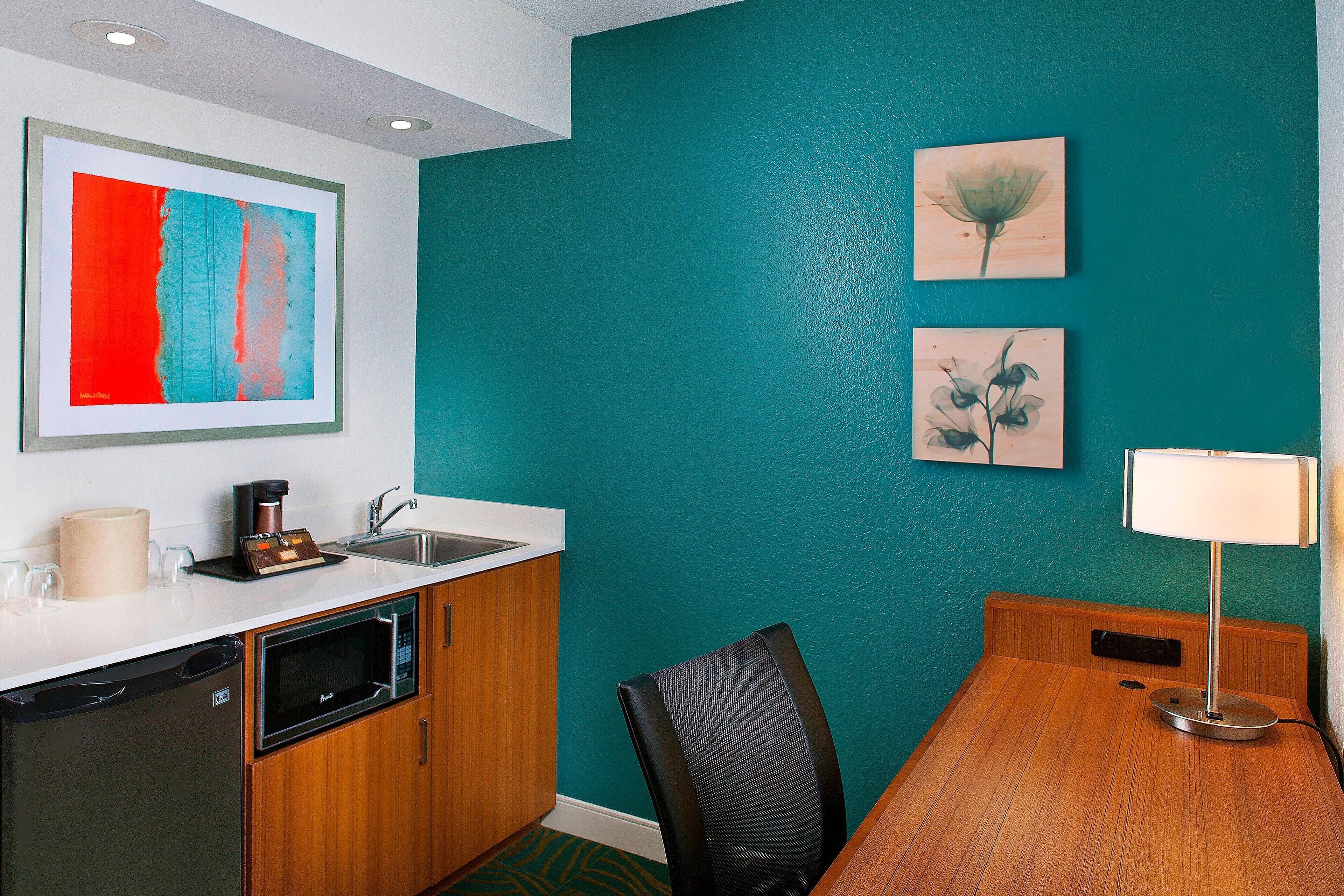 A compact kitchen with a microwave, coffee maker, and sink. There is a wooden dining table with a chair and a lamp. Two framed artworks are on the teal wall.