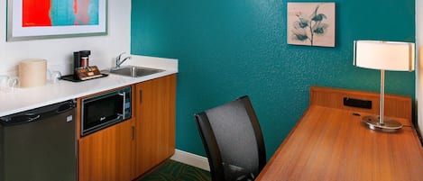 A compact kitchen with a microwave, coffee maker, and sink. There is a wooden dining table with a chair and a lamp. Two framed artworks are on the teal wall.