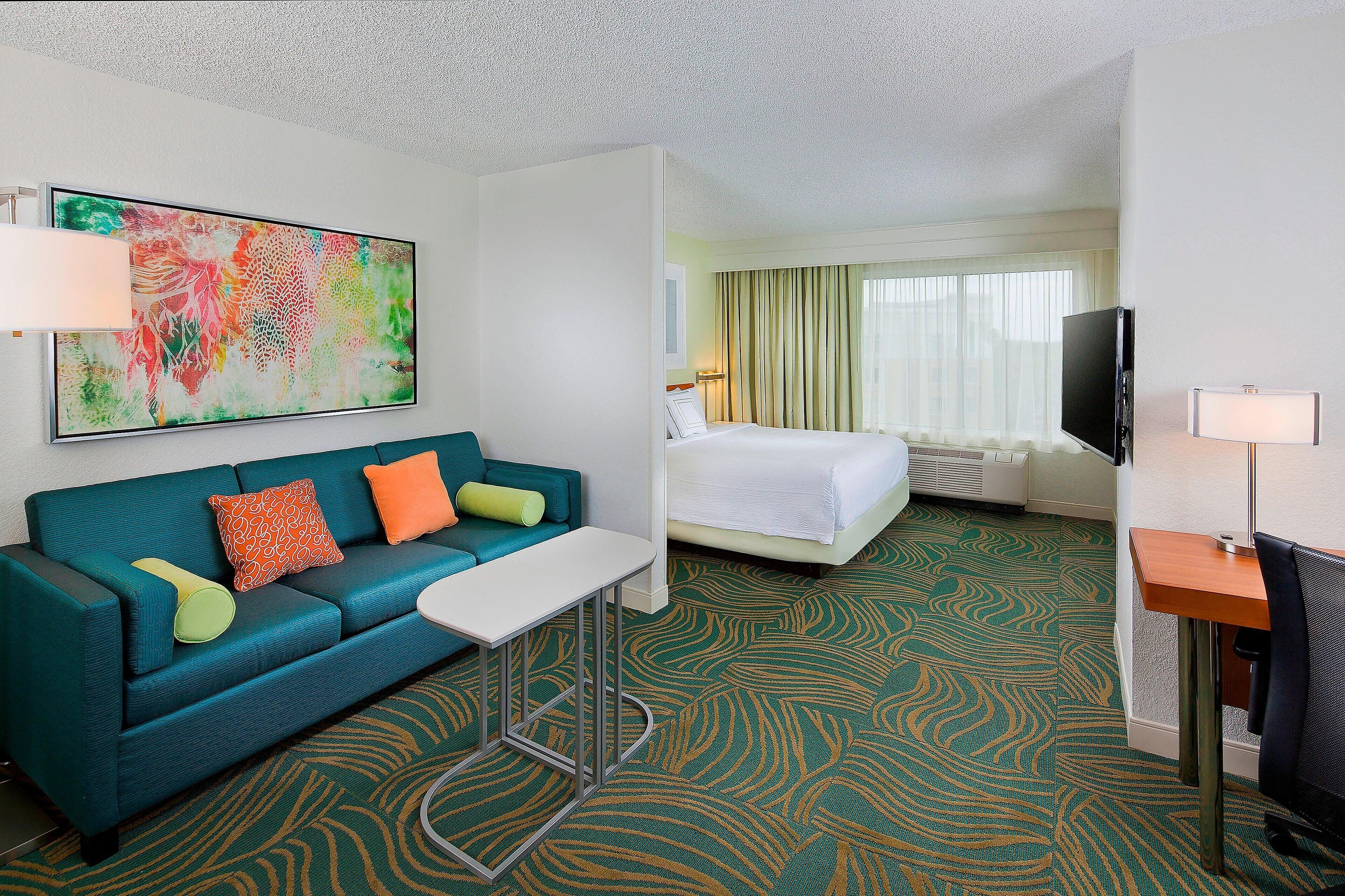A hotel room with a teal sofa, a small table, a bed, a TV, and a large abstract painting on the wall.