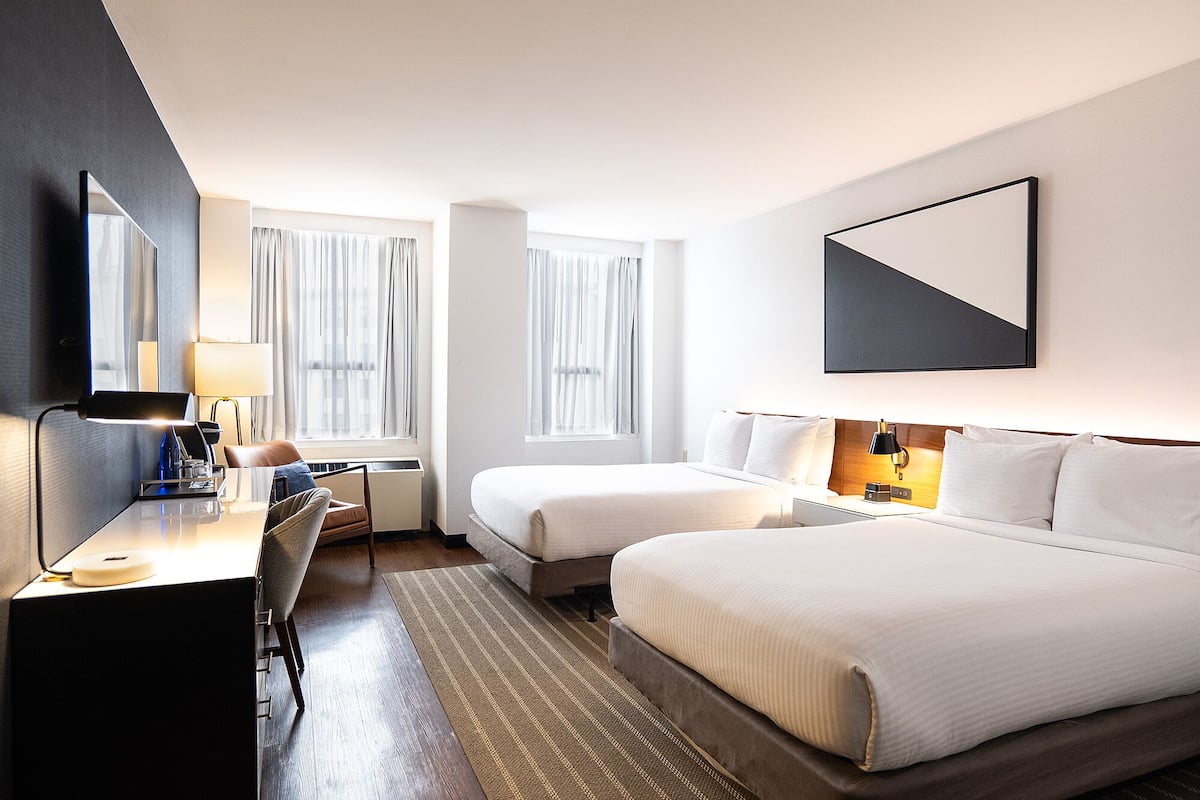 A hotel room with a desk, two beds, a large mirror, and a view of the city through the windows.