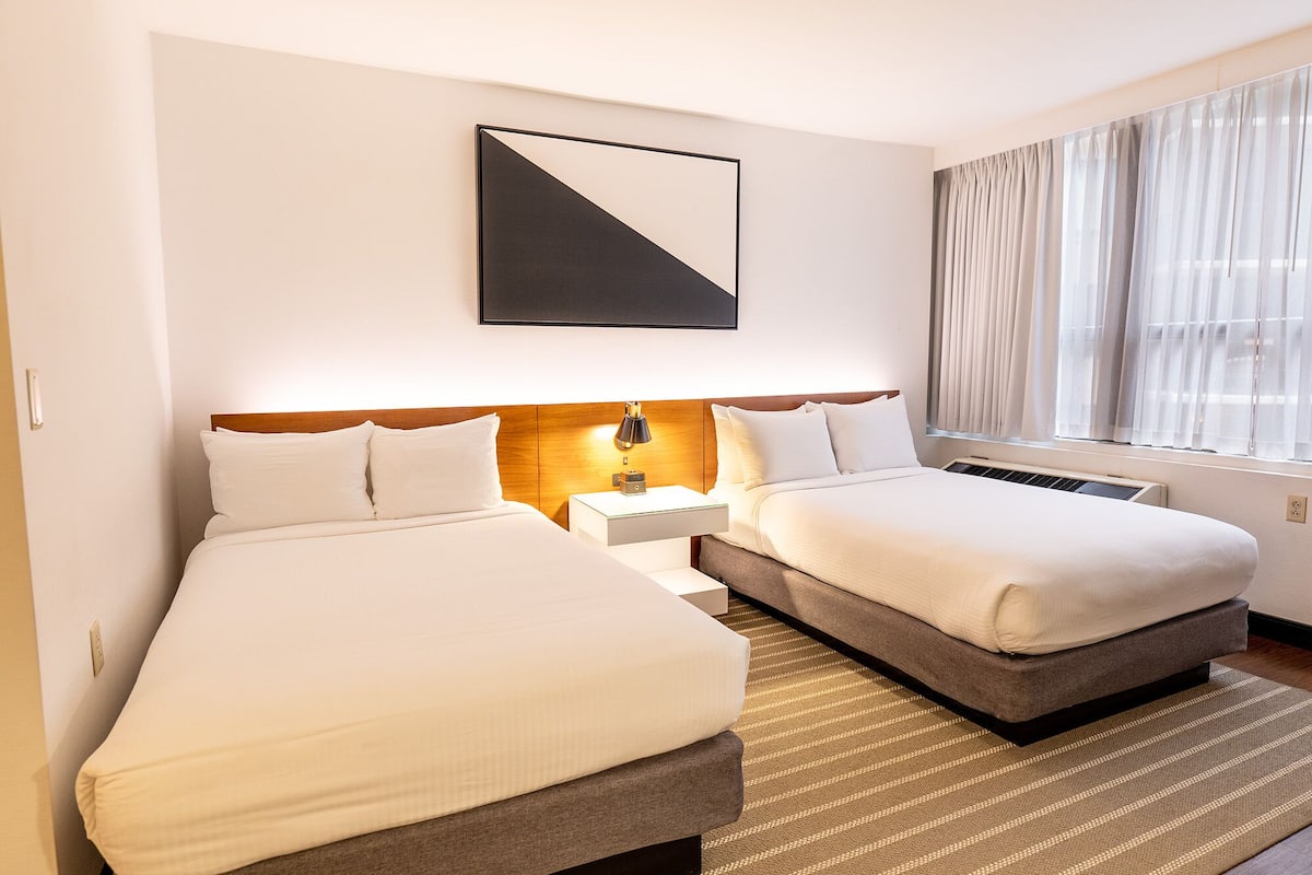 A hotel room with two beds, a modern headboard, a bedside table with a lamp, a large window with sheer curtains, and a geometric artwork on the wall.