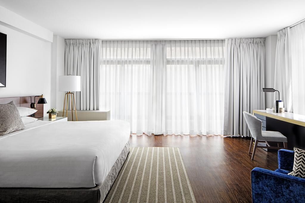 Habitación de hotel con cama, escritorio, silla y ventanas grandes con cortinas.