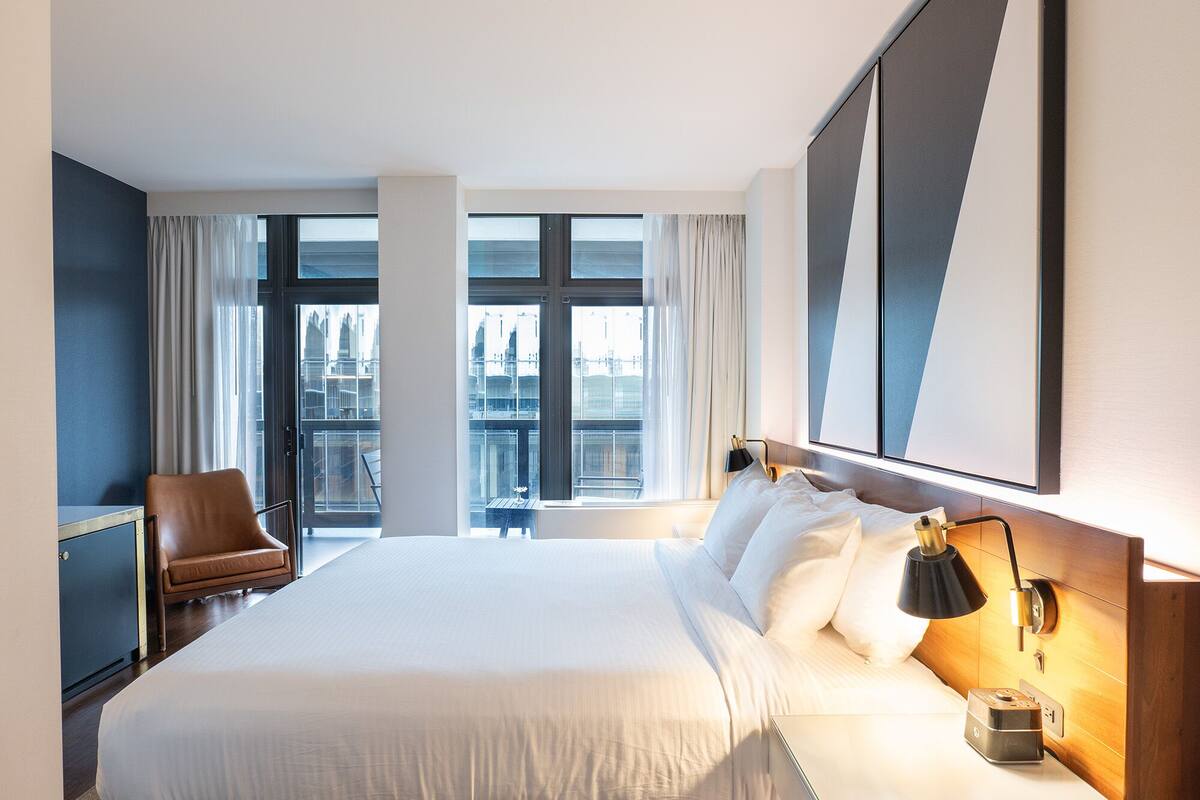 A hotel room with a large bed, a desk, a chair, and a view of the city through large windows.