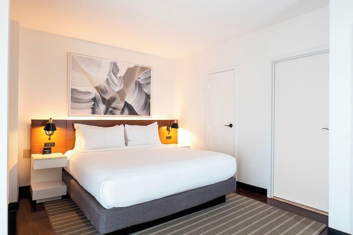 A hotel room with a large bed, two bedside tables with lamps, a wall-mounted artwork, and a closed door.