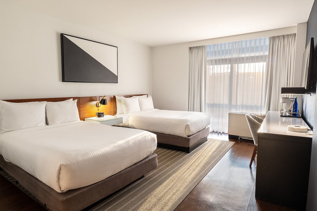 A hotel room with two beds, a desk, a chair, a TV, and a large window with curtains.