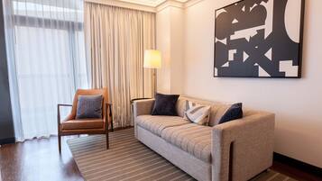 Quarto de hotel com sofá, cadeira, luminária e uma grande pintura abstrata na parede.