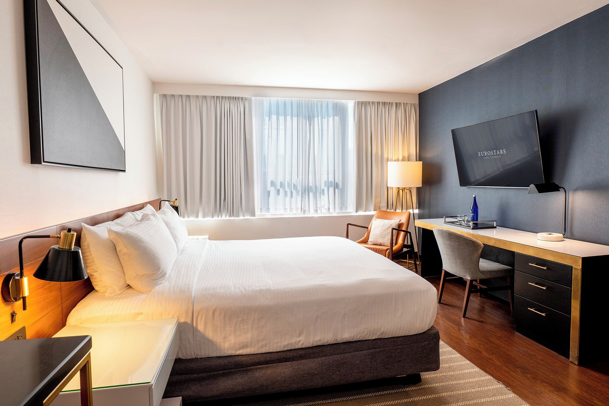Una habitación de hotel con cama, escritorio, silla, televisor y cortinas.