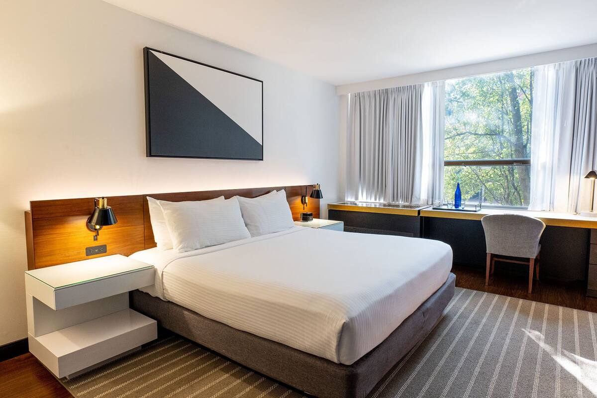 A modern hotel room with a large bed, bedside tables, a desk, and a window with curtains.