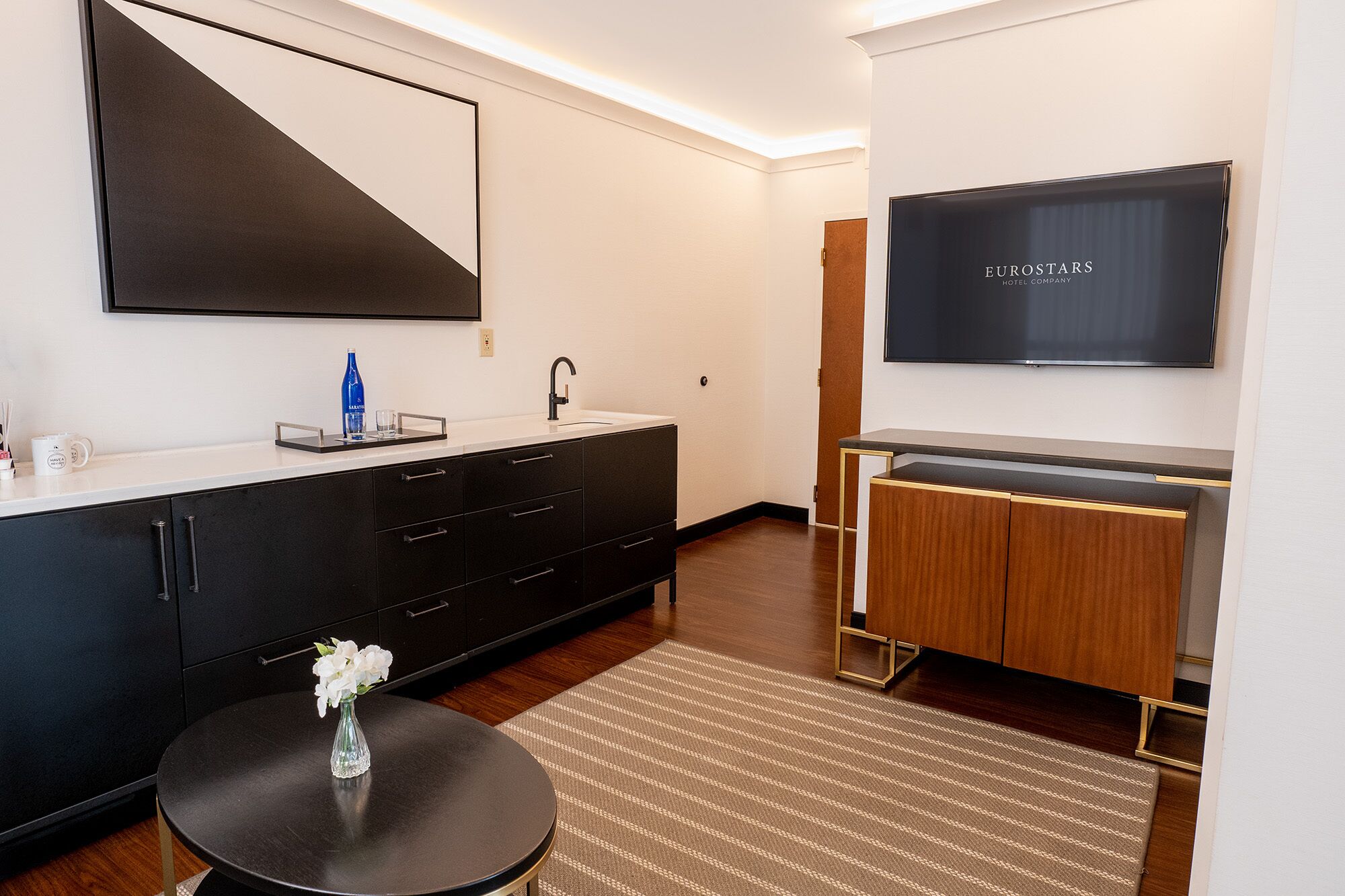 Une chambre d’hôtel moderne dotée d’un grand téléviseur à écran plat, d’un minibar noir et blanc, d’une table ronde agrémentée d’un vase de fleurs et d’un tapis à motifs rayés.