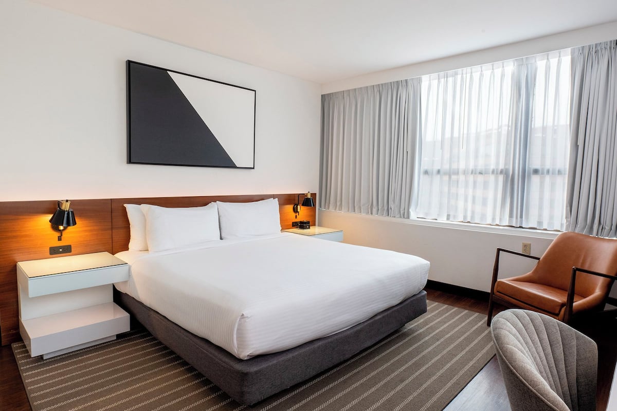 A hotel room with a bed, bedside tables, a chair, a window with curtains, and a wall-mounted picture.