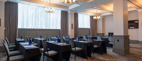 Un salón de conferencias con varias mesas y sillas, ventanas grandes con cortinas y un candelabro.