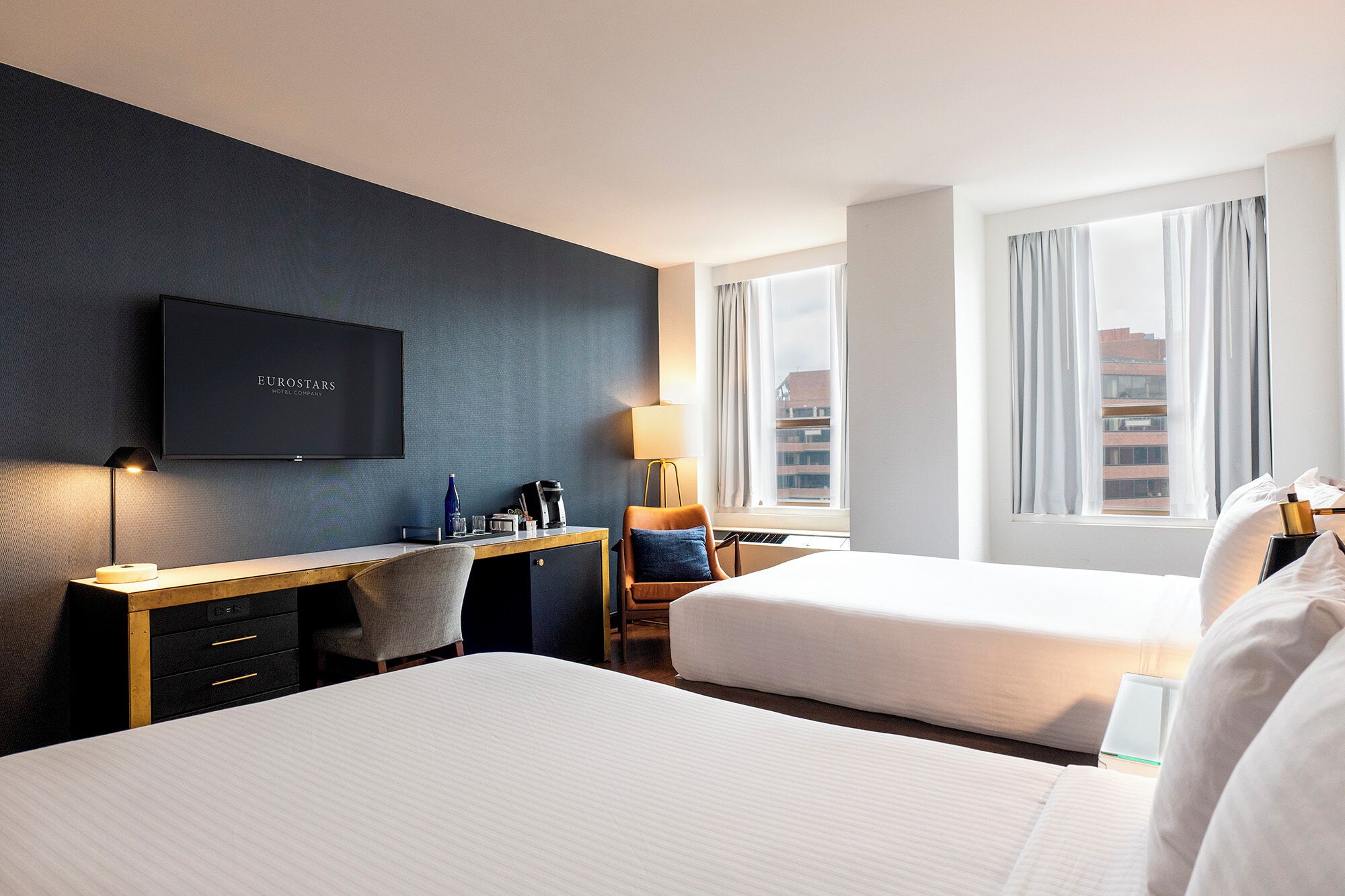A hotel room with a dark blue accent wall, a flat-screen TV, a desk with a chair, a bed, and a window with curtains.