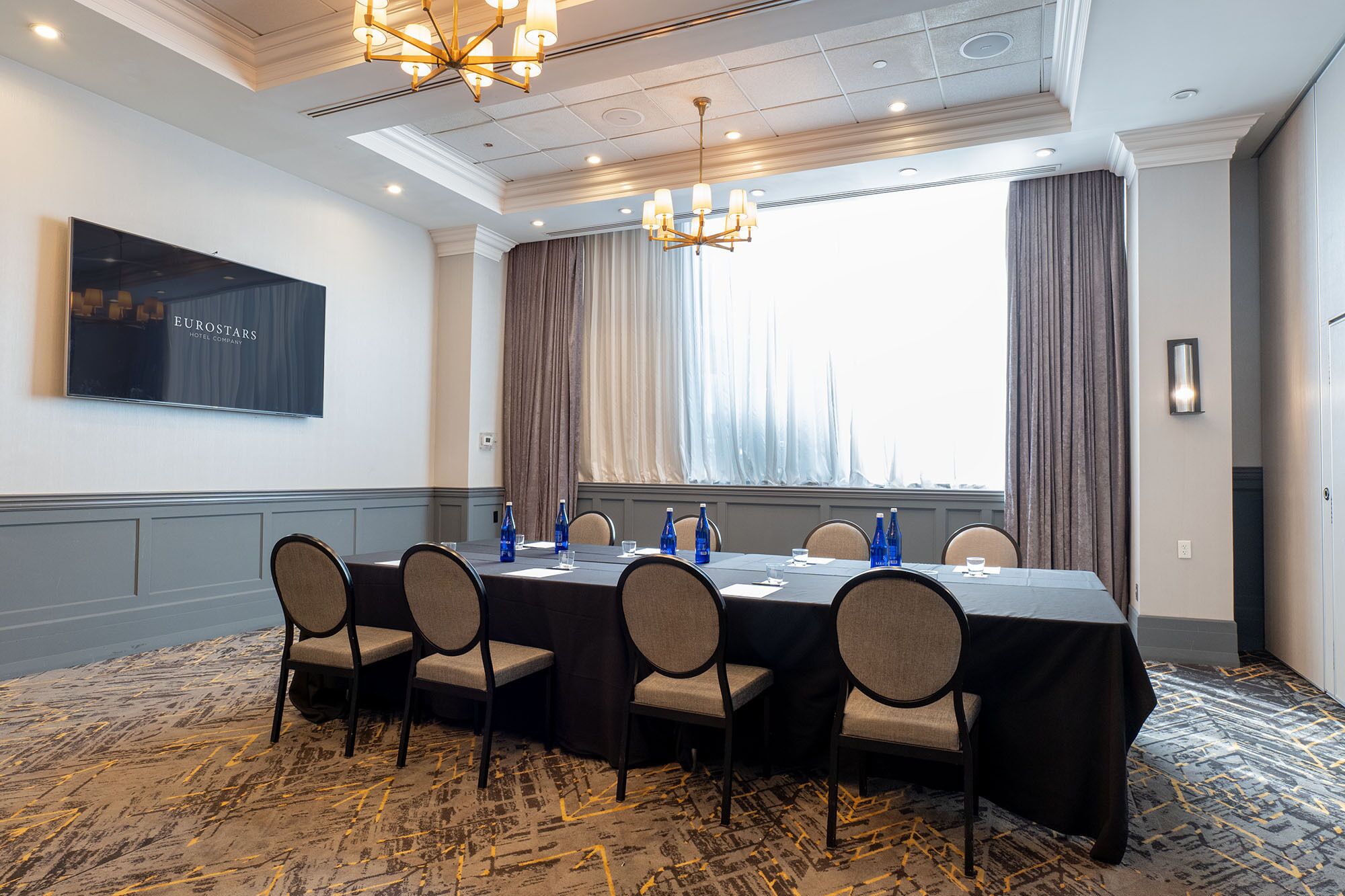 Uma sala de conferência com uma mesa comprida, cadeiras e uma grande janela com cortinas.