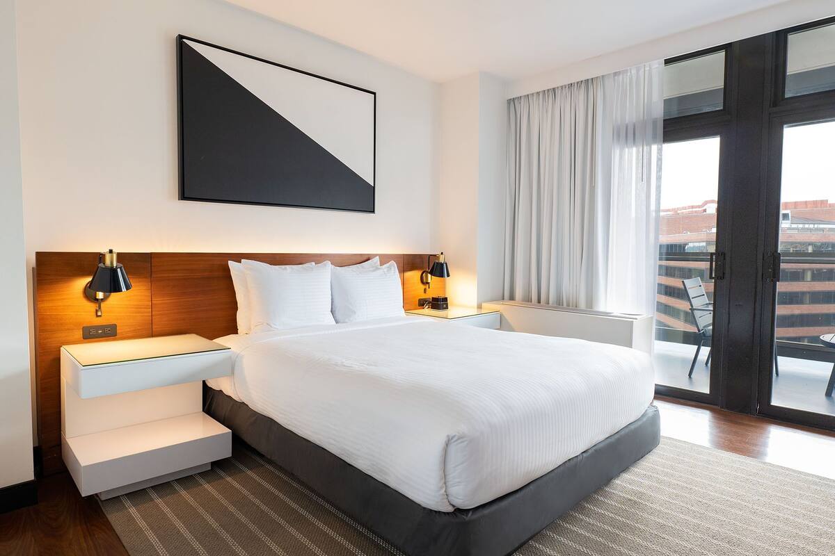A hotel room with a large bed, two bedside tables with lamps, a large window with curtains, and a city view.