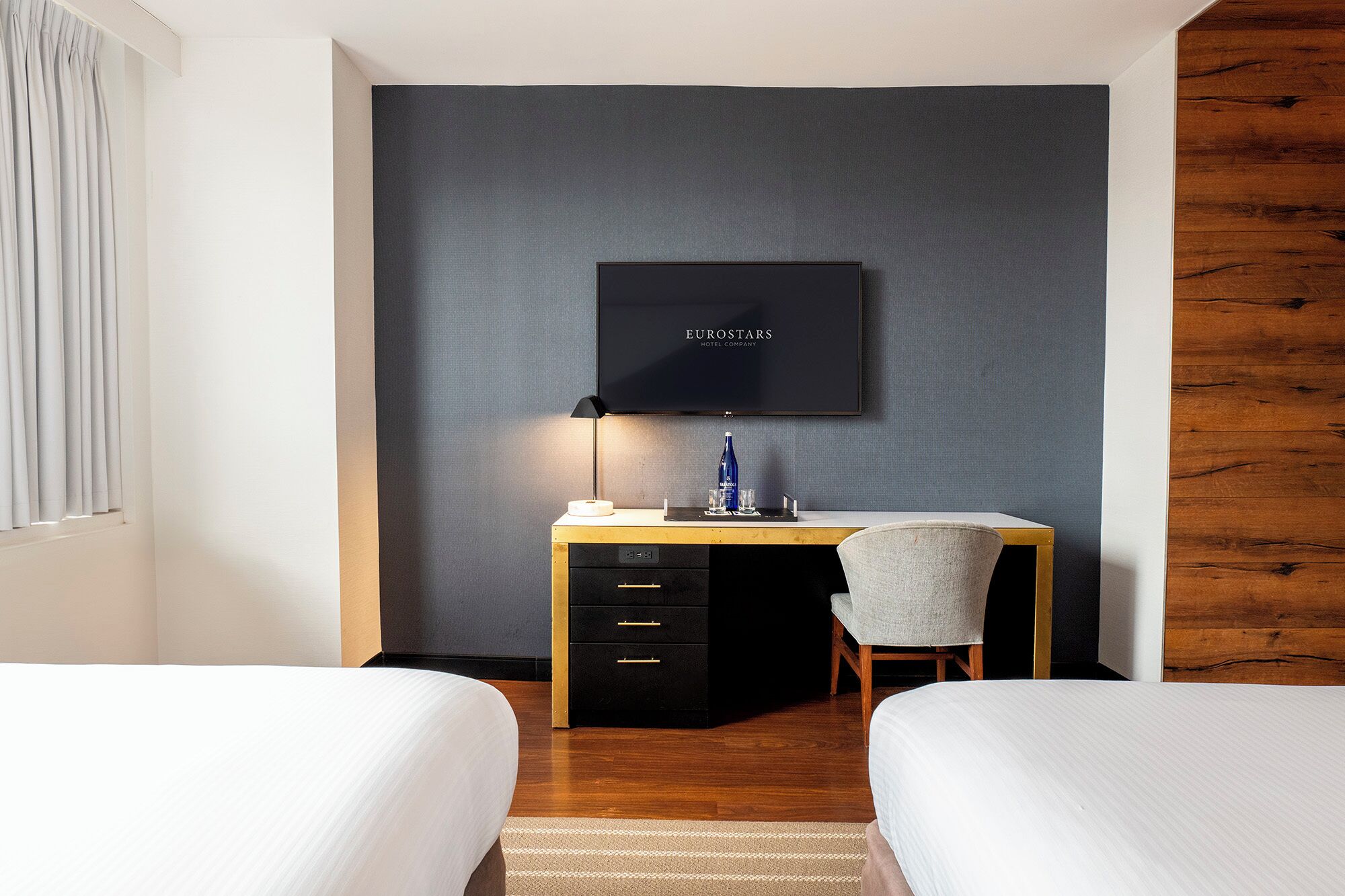 A hotel room with a bed, a desk with a chair, a television mounted on the wall, and a wooden accent wall.