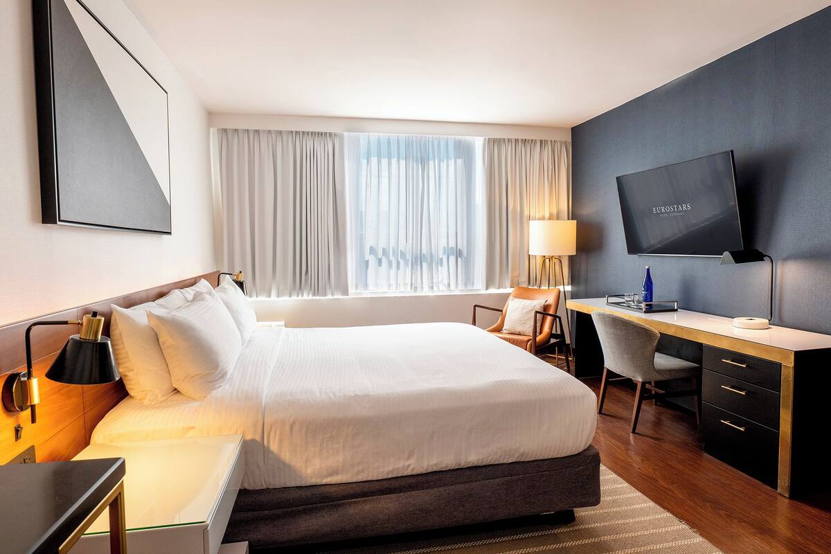 A hotel room with a bed, desk, chair, TV, and curtains.
