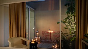 Habitación de hotel con cama, una silla, una mesita con velas y vistas a un balcón con plantas y barandilla.