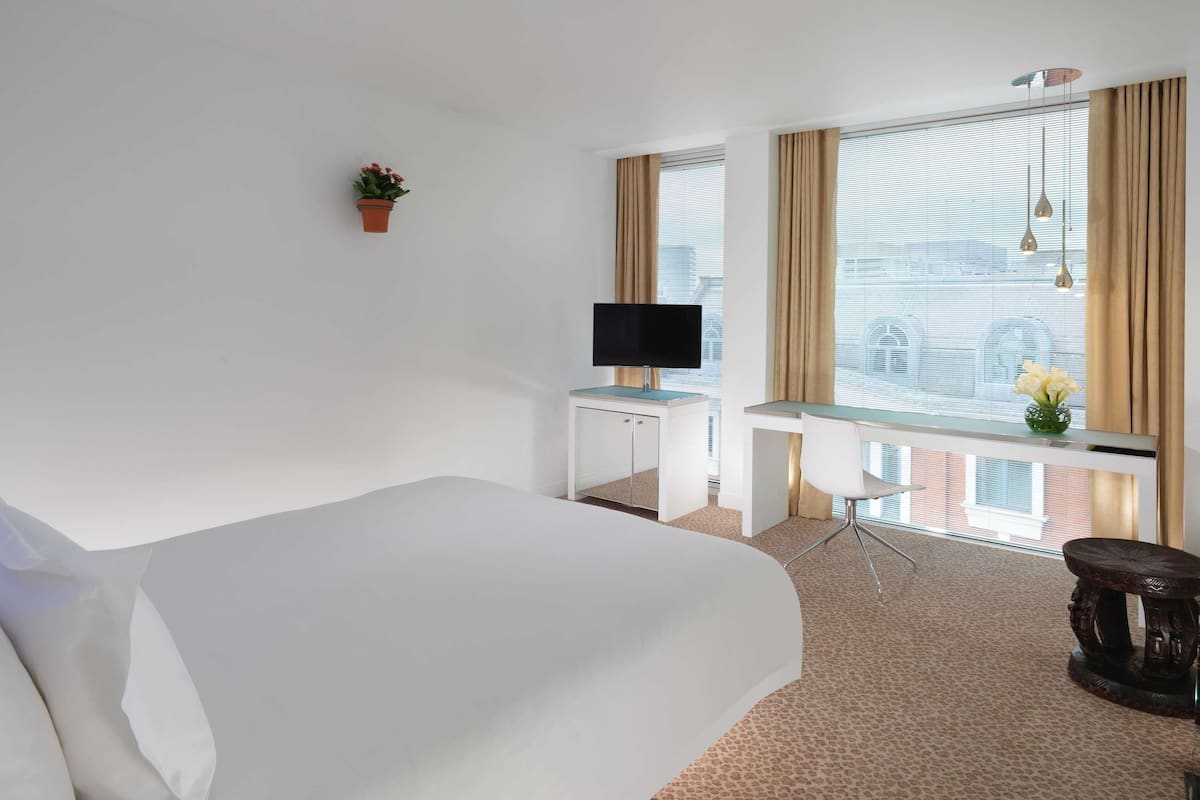 Ein Hotelzimmer mit Bett, Schreibtisch, Fernseher, einem Fenster mit Vorhängen und einer kleinen Pflanze.