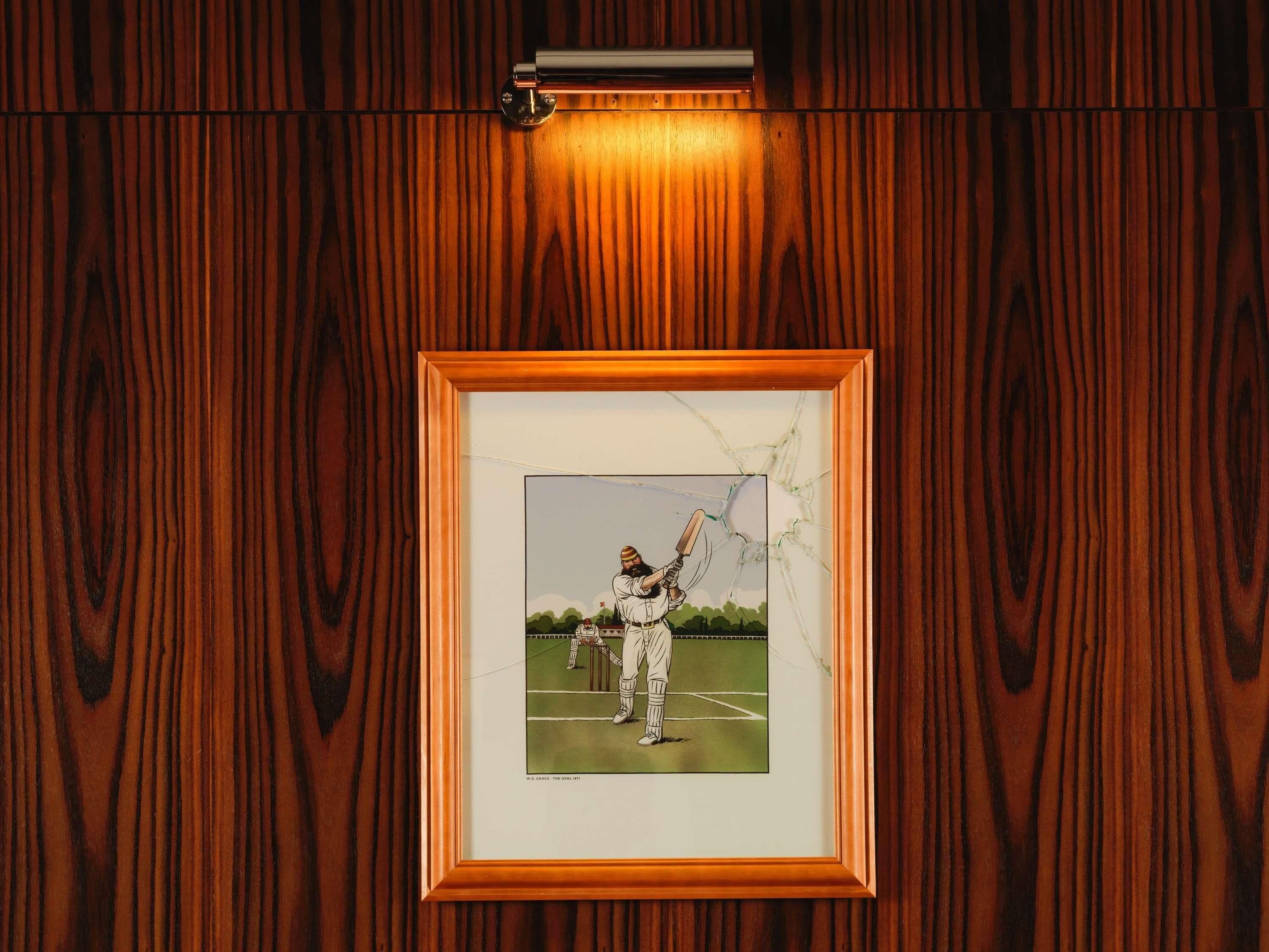 A framed picture of a cricket player on a field, mounted on a wooden wall.