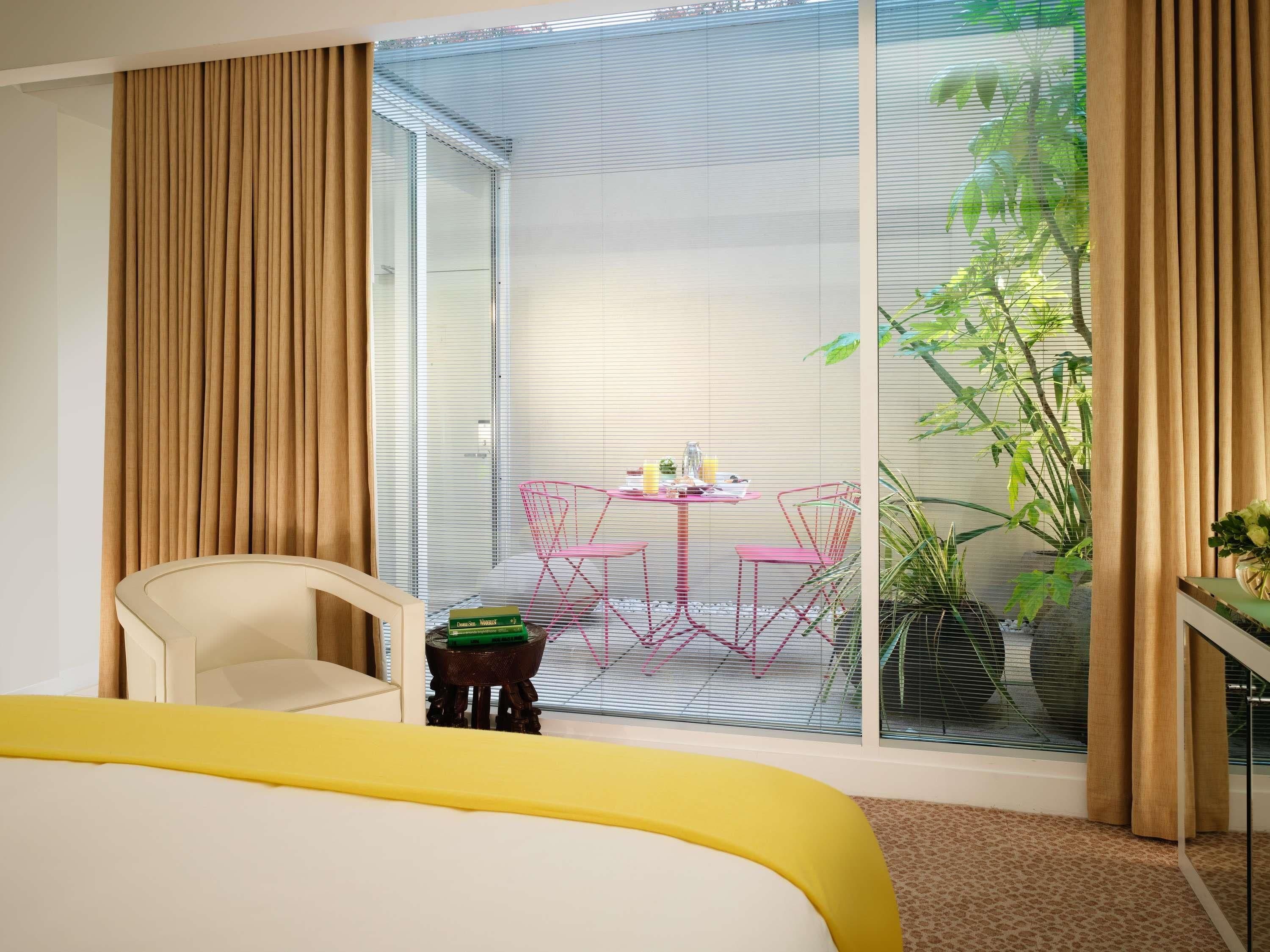 Habitación de hotel con cama, una pequeña mesa con sillas, una ventana con persianas y una planta en maceta.