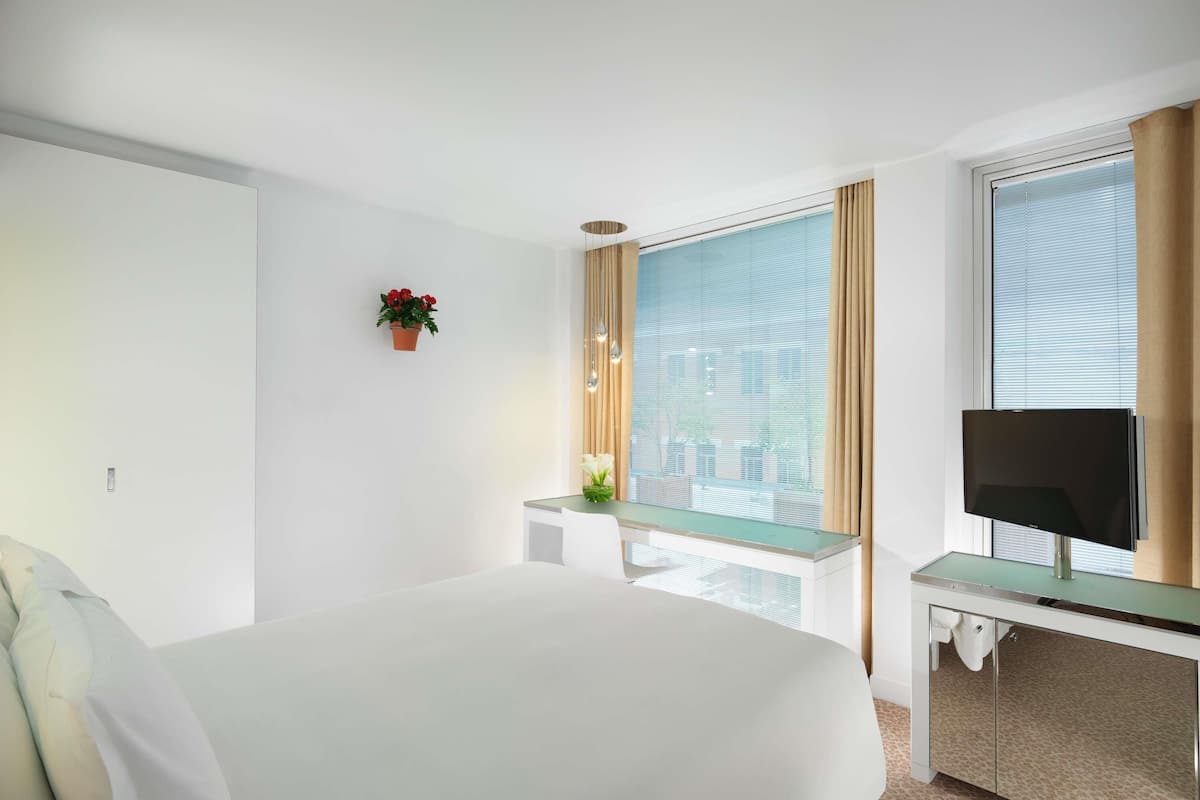 Ein Hotelzimmer mit Bett, kleinem Schreibtisch mit Fernseher, Fenster mit Vorhängen und einer Zimmerpflanze.