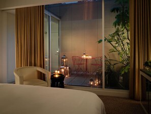 A hotel room with a bed, a chair, a small table with candles, and a view of a red chair and plants through a window. - St Martins Lane London, a Morgans Originals hotel (London)