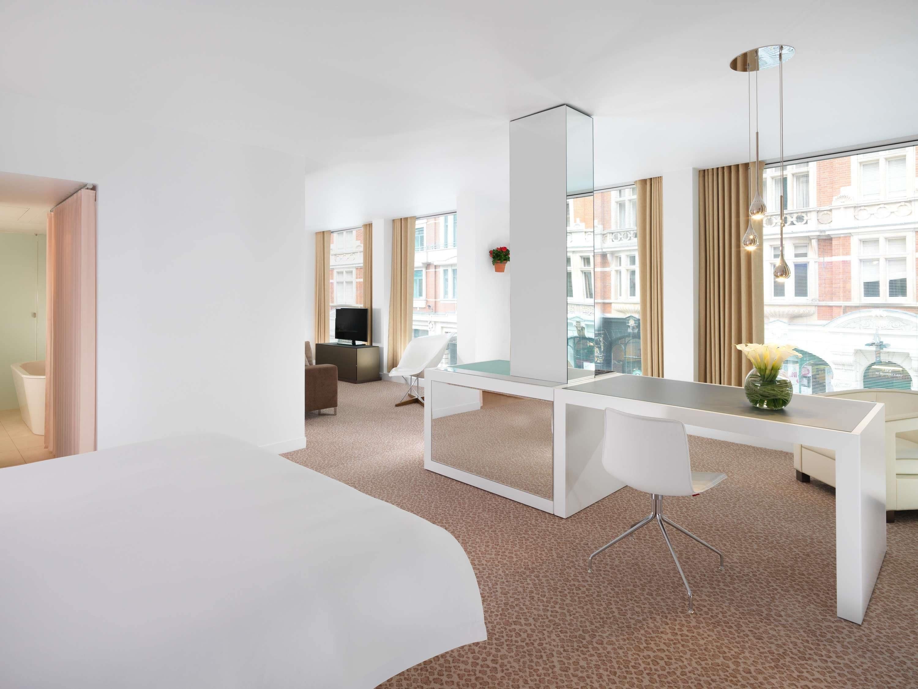 A modern hotel room with a large bed, a desk with a chair, a television, and large windows with curtains.