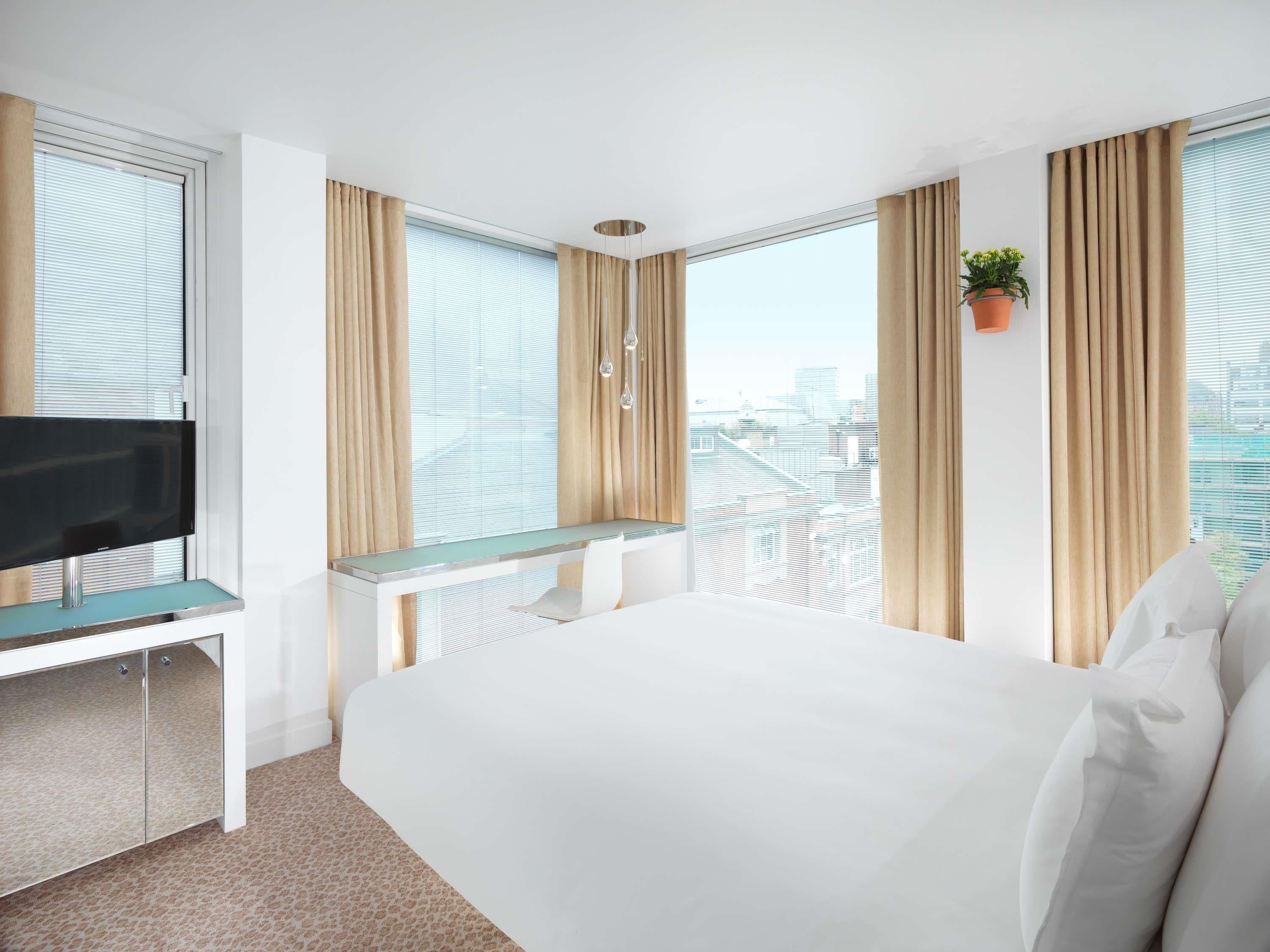 A hotel room with a bed, a desk with a TV, and large windows with curtains.